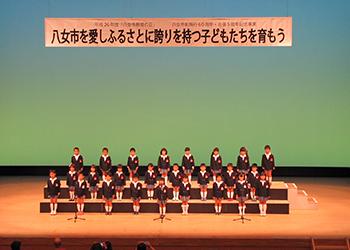 「八女市を愛しふるさとに誇りを持つ子どもたちを育もう」と書かれた舞台の横断幕の下に子供達が並んでいる写真