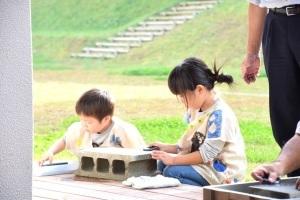 収穫体験に参加した女の子と男の子が、ブロックを台にして石庖丁を削っている写真