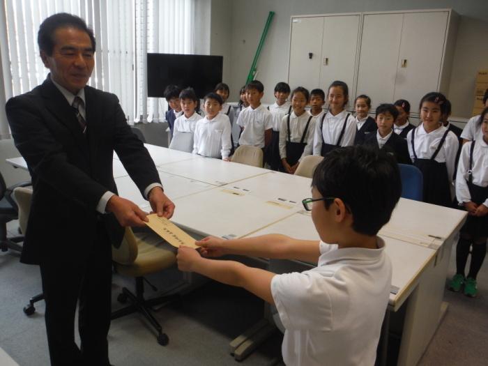 井上立花支所長へ寄付金を手渡している筑南小学校の代表の生徒とそれを見守っている他の生徒の写真
