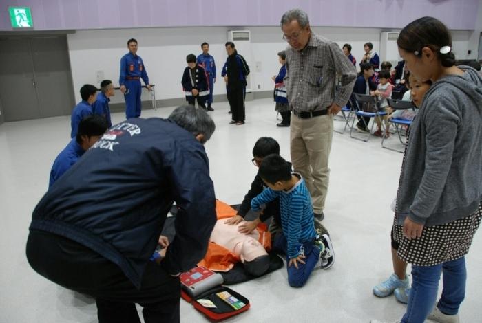 マネキンの横にAEDの機械が入った袋を広げて、消防士の説明を聞きながら年配の男性や子ども達が心肺蘇生法を教わっている写真