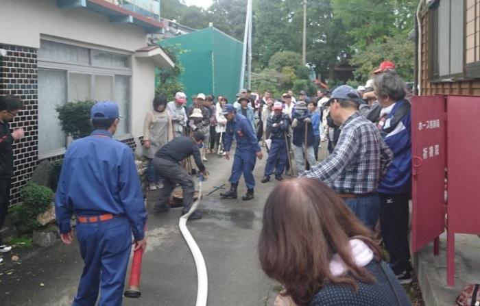 参加者や消防士が建物の外に出て、男性が白いホースを伸ばして水を出し、火事がおきた場合の訓練を行っている写真