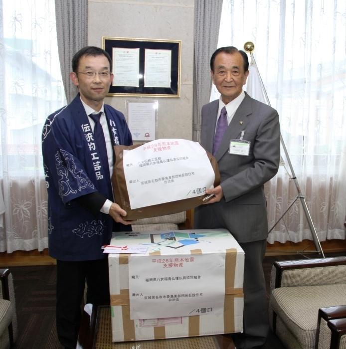 伝統的工芸品と書かれたハッピを着たメガネ姿の男性が箱に入った支援物資を市長に手渡している写真