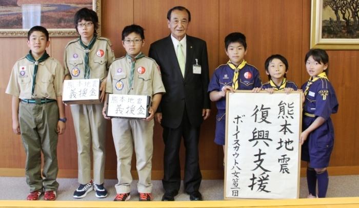 真ん中に市長、右となりには熊本地震復興支援ボーイスカウト八女第一団と書かれた案内をもつ青い衣装の子ども3人、左隣には募金箱を持ちたっている薄緑の衣装の男の子3人の写真