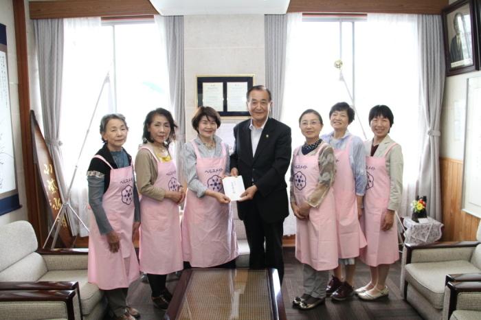 ピンクのお揃いのエプロン姿の八女市食生活改善推進会の女性6名が笑顔で市長に義援金を手渡している写真