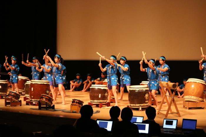 舞台で星野小学校の生徒達がハッピ姿で大、中、小の太鼓をばちでたたいている写真