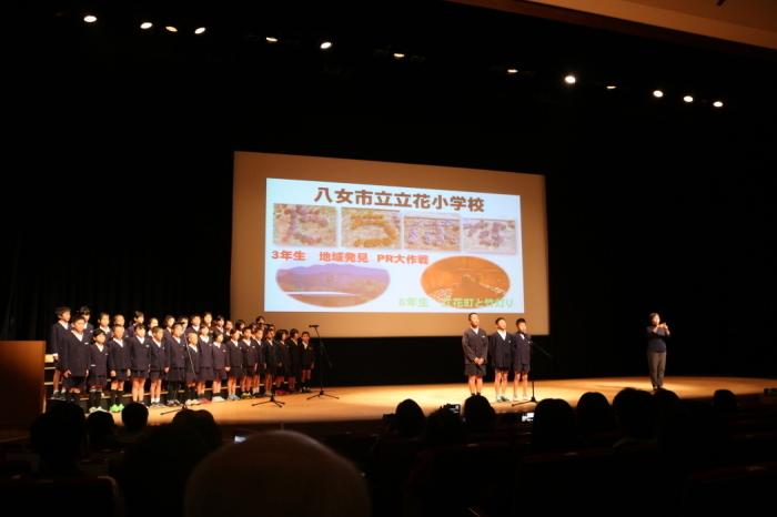 立花町小学校の生徒3人が舞台中央のマイクの前にたち、他の生徒は舞台左横に並んで発表をしている写真