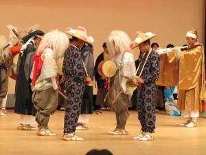 舞台中央で向き合いながら立っている笠を被った男性、白髪姿で太鼓を抱えている人などの写真