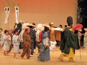 黄色と緑色の袈裟を着ている人を先頭に袴姿の少年、浴衣姿の子供達が後に続いて並んで歩いている劇の写真