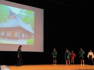 八女ふる里学発表の舞台の大きなスクリーンに家が映し出されて、その前で演技をしている子供達の写真