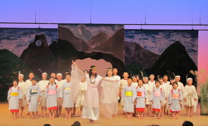 八女津媛伝説の劇で白い衣装をきた女の子が踊っている後方で他の役柄の生徒達が歌をうたっている写真