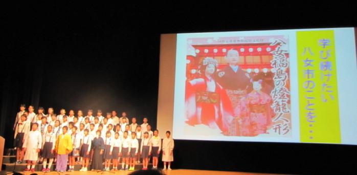 舞台右に大きなモニターが写り、その横で福島小学校の生徒達が発表している写真