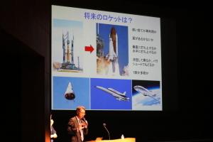 舞台上のスクリーンにロケットモニターが映し出され、中須賀真一氏が講演をしている写真