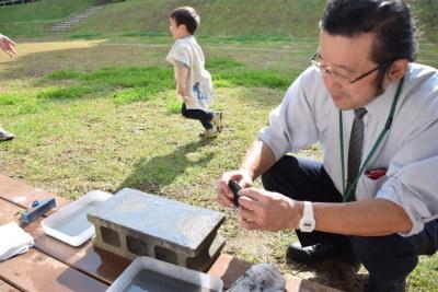 石庖丁づくりに飽きた男の子が後ろで遊んでいるのを、シャツにネクタイを締めた男性が変わり作っている写真