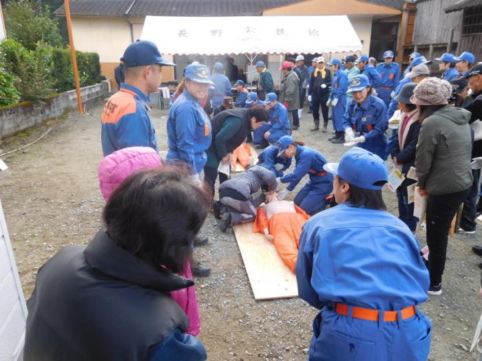 公民館の広場で、人が乗るくらいの木の板の上にマネキンを横たわらせ、女性の消防団の指導のもと参加者の市民がマネキンの顔に近づけて訓練を行っている様子を消防団や市民の女性たちが見ている写真