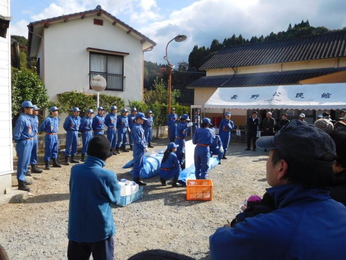 公民館の広場の中央で、女性の消防団が2つのビニールシートにくるまれたものに機材を用いて上げようとしている訓練を、横に整列した消防団や訓練に参加した市民が見ている写真